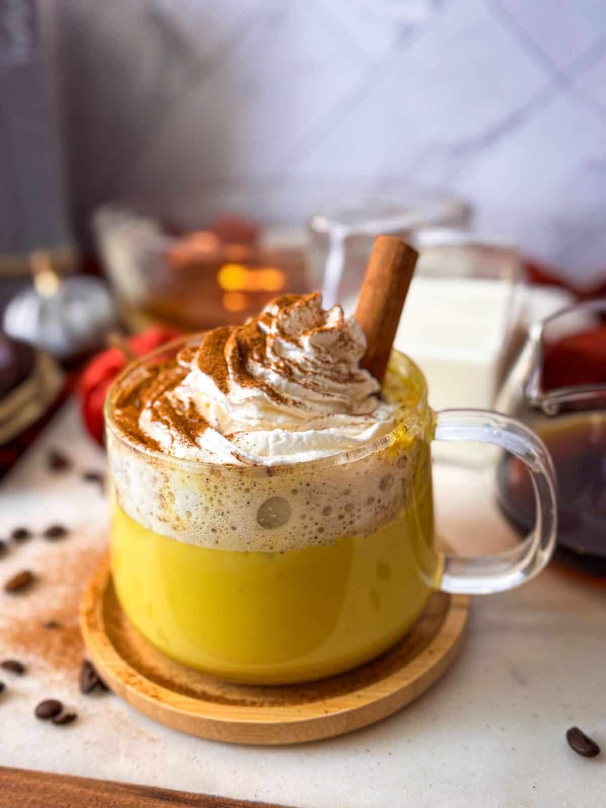 Banana Date Golden Milk Coffee Latte A glass mug filled with a creamy golden milk banana date coffee latte, topped with a swirl of whipped cream, a dusting of cinnamon, and a cinnamon stick, set on a wooden coaster.