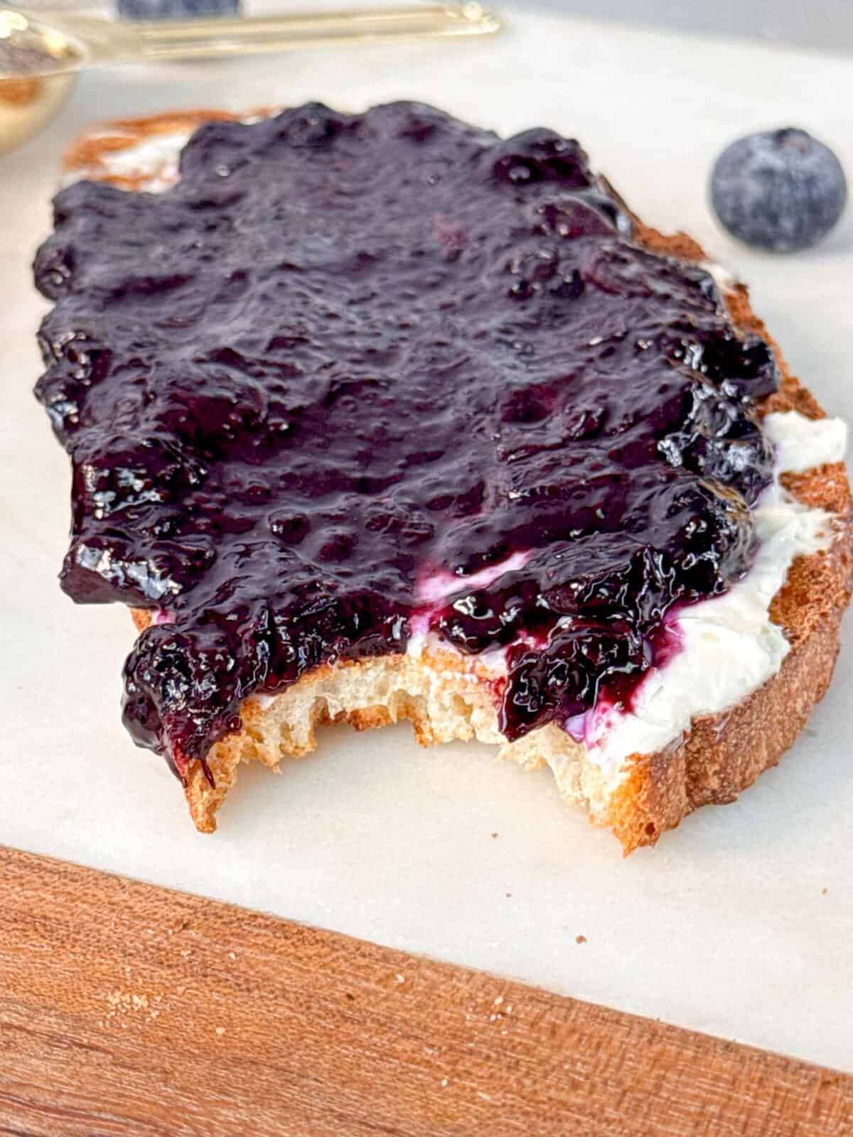 Blueberry Chia Jam Recipe A slice of bread topped with cream cheese and blueberry chia jam, with a bite taken out.
