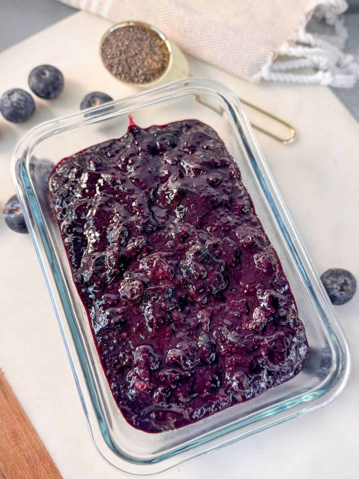 Blueberry Chia Seed Jam A glass container filled with homemade blueberry chia seed jam, surrounded by fresh blueberries.