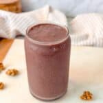 A close up shot of a glass filled with a blueberry date smoothie on a marble surface, surrounded by small granola clusters.