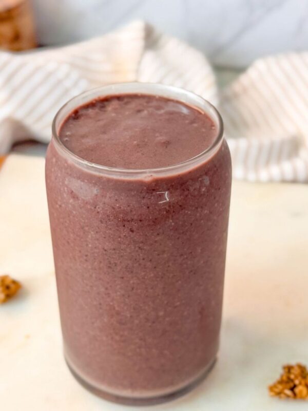 A close up shot of a glass filled with a blueberry date smoothie on a marble surface, surrounded by small granola clusters.