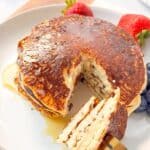 A close up shot of a fork cutting into a stack of cottage cheese almond flour pancakes drizzled with syrup, with strawberries and blueberries on the side.