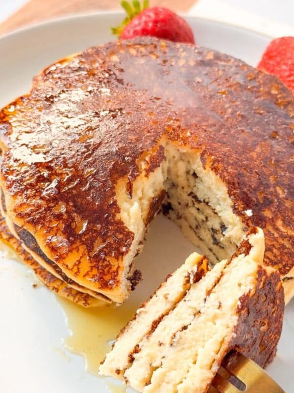 A close up shot of a fork cutting into a stack of cottage cheese almond flour pancakes drizzled with syrup, with strawberries and blueberries on the side.