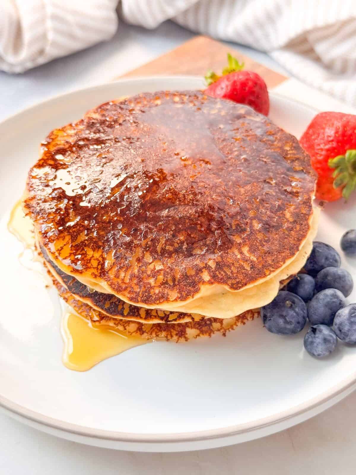 Fluffy Cottage Cheese Almond Flour Pancakes A stack of almond flour cottage cheese pancakes topped with syrup, served with fresh strawberries and blueberries on a white plate.