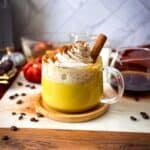A glass mug filled with a creamy golden milk banana date coffee latte, topped with a swirl of whipped cream, a dusting of cinnamon, and a cinnamon stick, surrounded by coffee beans and autumn decor.