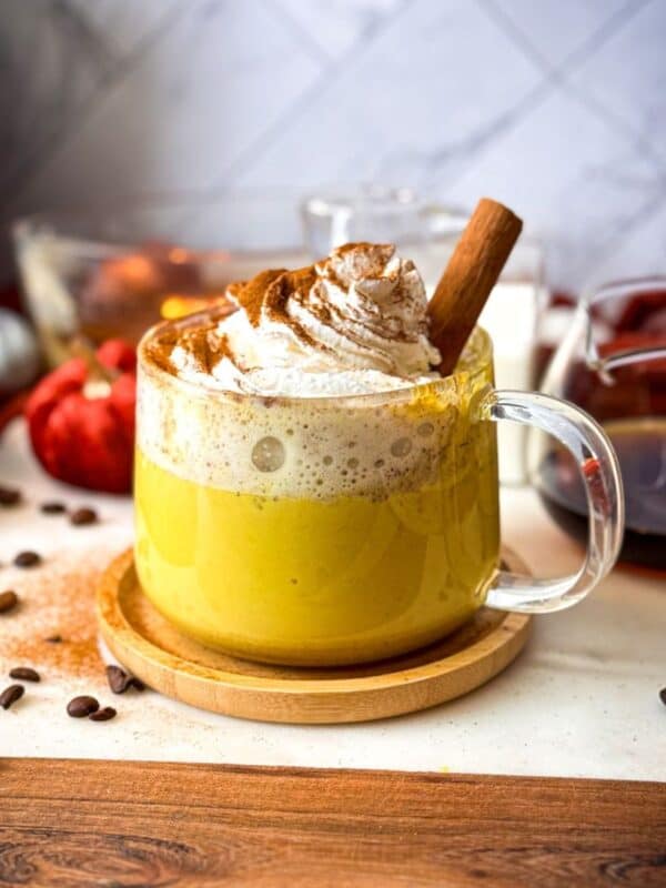 A glass mug filled with a creamy golden milk banana date coffee latte, topped with a swirl of whipped cream, a dusting of cinnamon, and a cinnamon stick, surrounded by coffee beans and autumn decor.