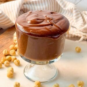 A glass cup filled with creamy homemade Nutella, surrounded by hazelnuts and rice cakes.