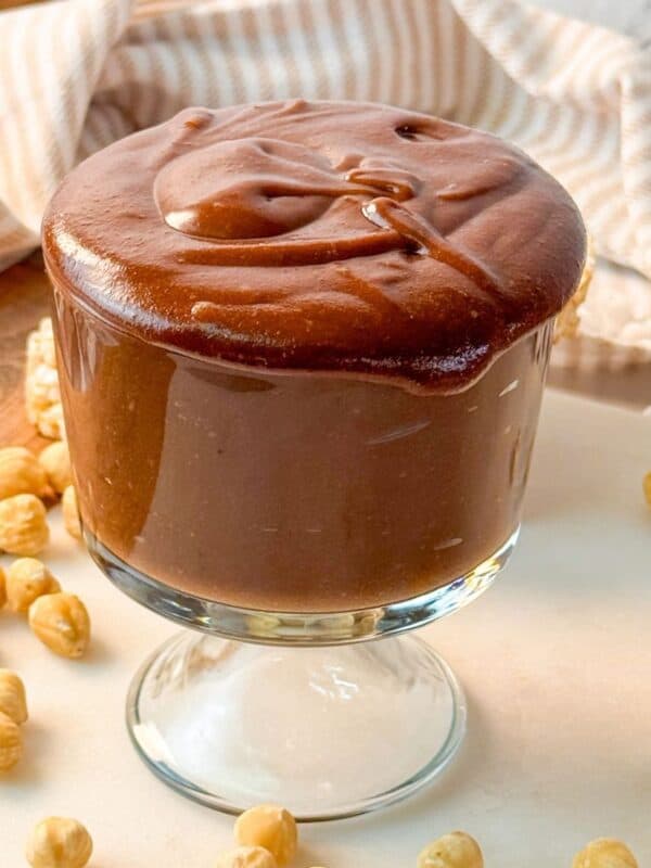 A glass cup filled with creamy homemade Nutella, surrounded by hazelnuts and rice cakes.