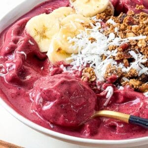 Spoonful of thick frozen berry smoothie bowl topped with banana slices, coconut, and granola