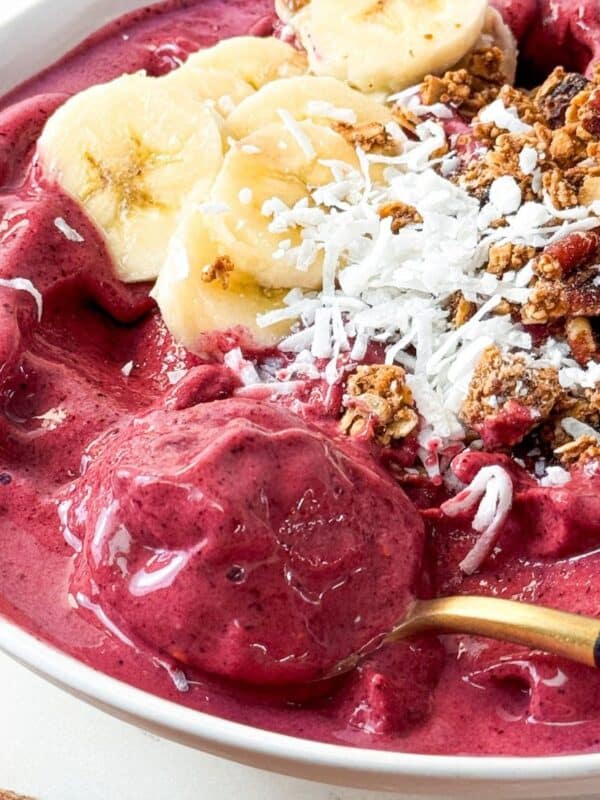 Spoonful of thick frozen berry smoothie bowl topped with banana slices, coconut, and granola