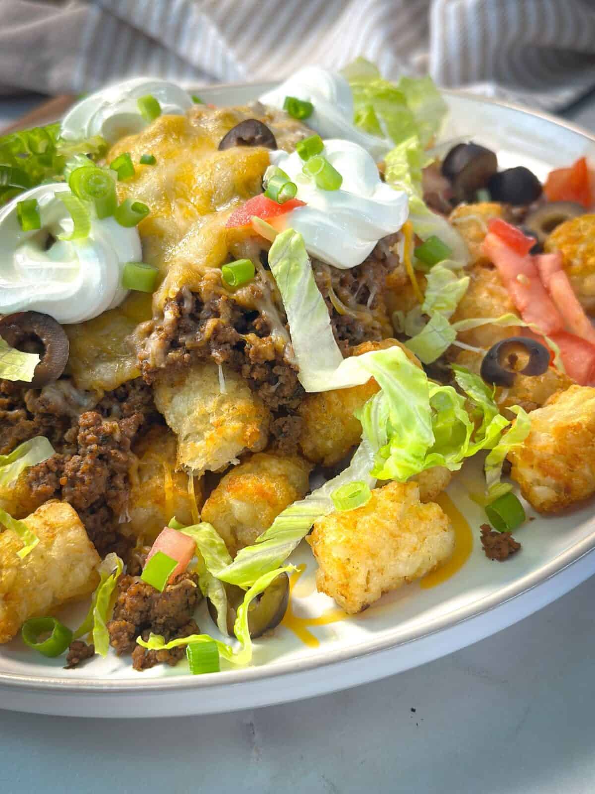 Taco Tater Tot Casserole A close-up shot of a tater taco tot casserole topped with seasoned ground beef, melted cheese, shredded lettuce, diced tomatoes, sliced black olives, green onions, and dollops of sour cream.