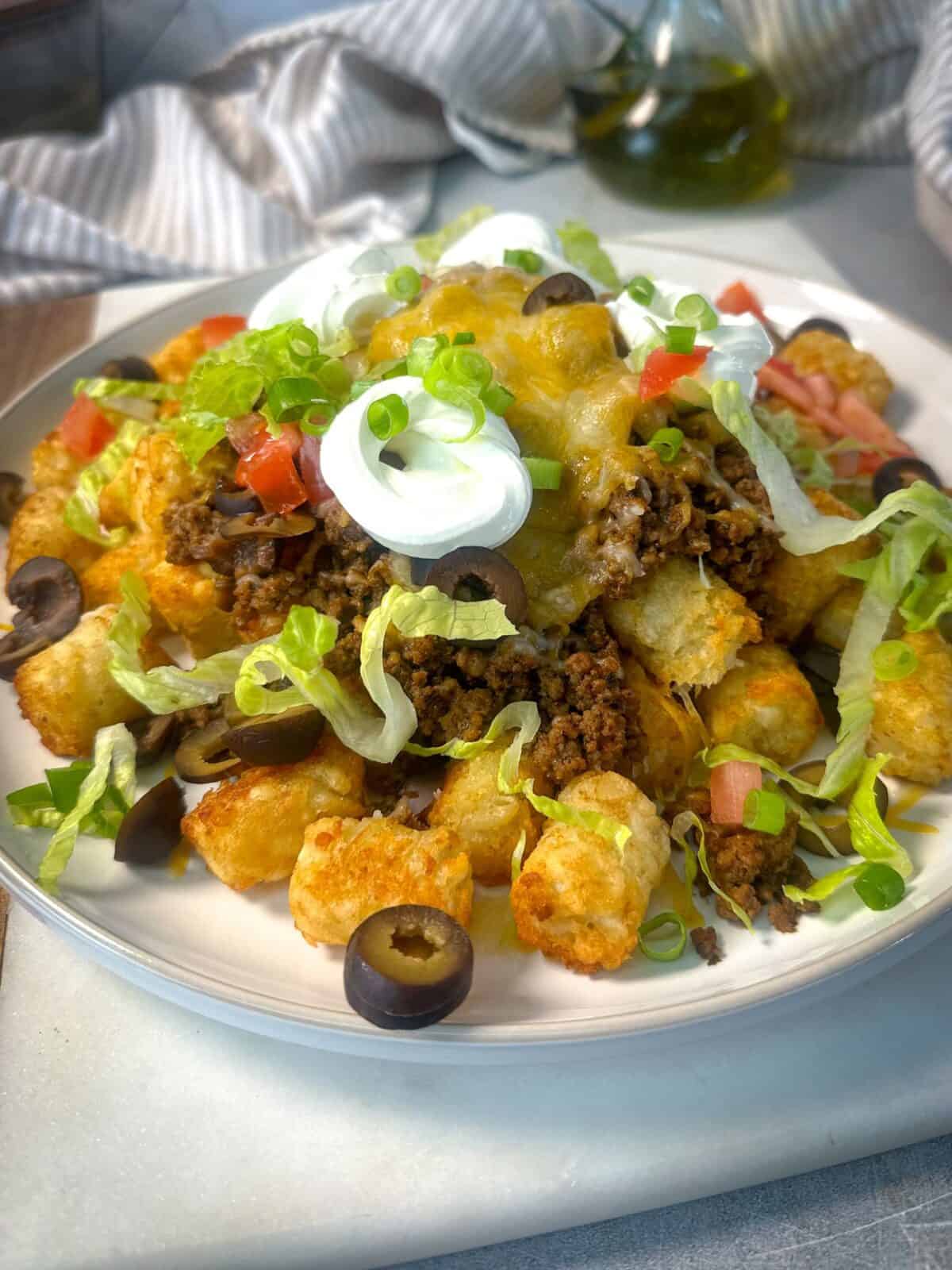 Taco Tater Tot Casserole Recipe A plate of loaded taco tater tot casserole with crispy tater tots, taco-seasoned beef, melted cheese, shredded lettuce, tomatoes, black olives, green onions, and sour cream, ready to be served.