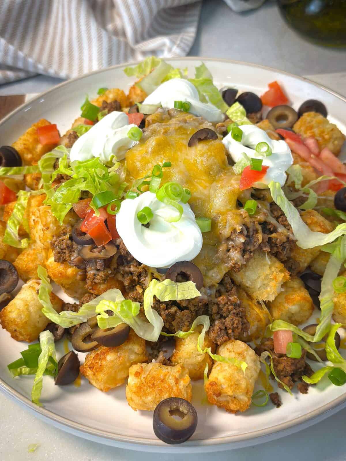 Tater Taco Tot Casserole A shot of a cheesy taco tater tot topped with seasoned ground beef, melted cheese, shredded lettuce, diced tomatoes, sliced black olives, green onions, and dollops of sour cream.