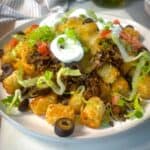 A close-up of a cheesy taco tater tot casserole topped with seasoned ground beef, melted cheese, shredded lettuce, diced tomatoes, sliced black olives, green onions, and dollops of sour cream.