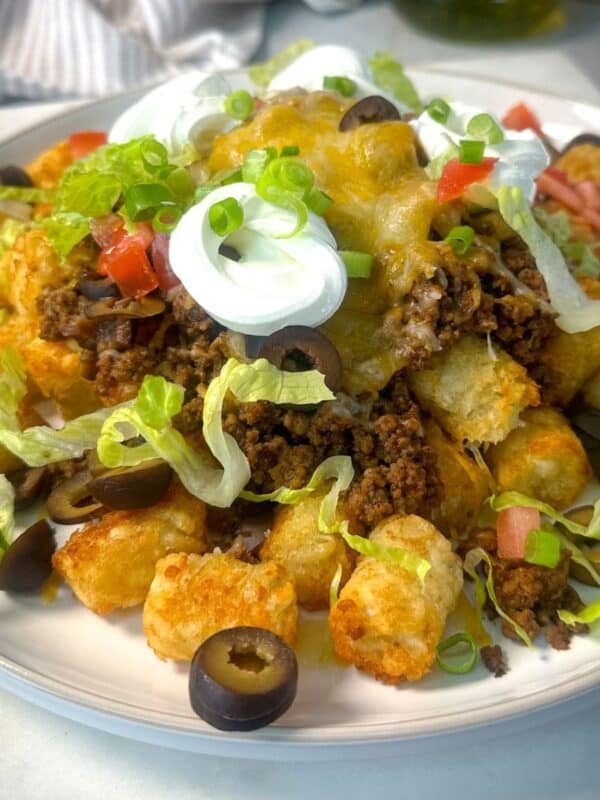 A close-up of a cheesy taco tater tot casserole topped with seasoned ground beef, melted cheese, shredded lettuce, diced tomatoes, sliced black olives, green onions, and dollops of sour cream.