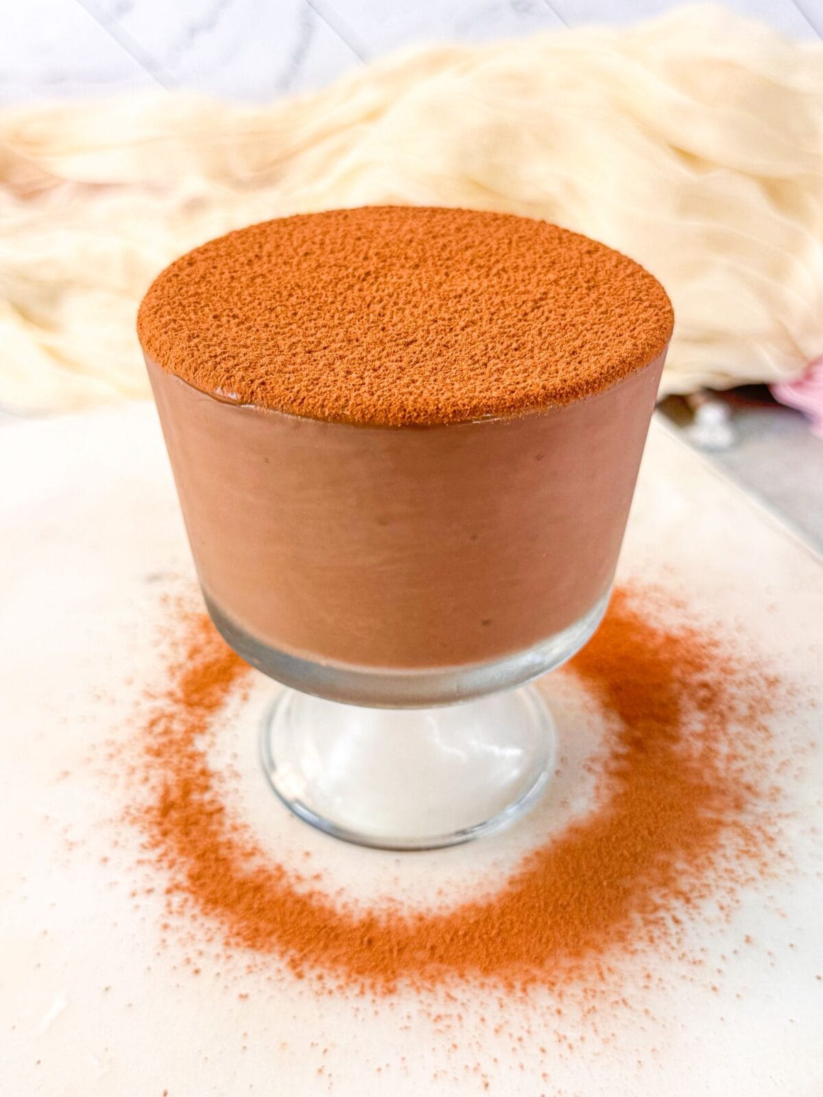 A chocolate protein pudding topped with cacao powder, surrounded by a light cocoa dusting on a white surface.