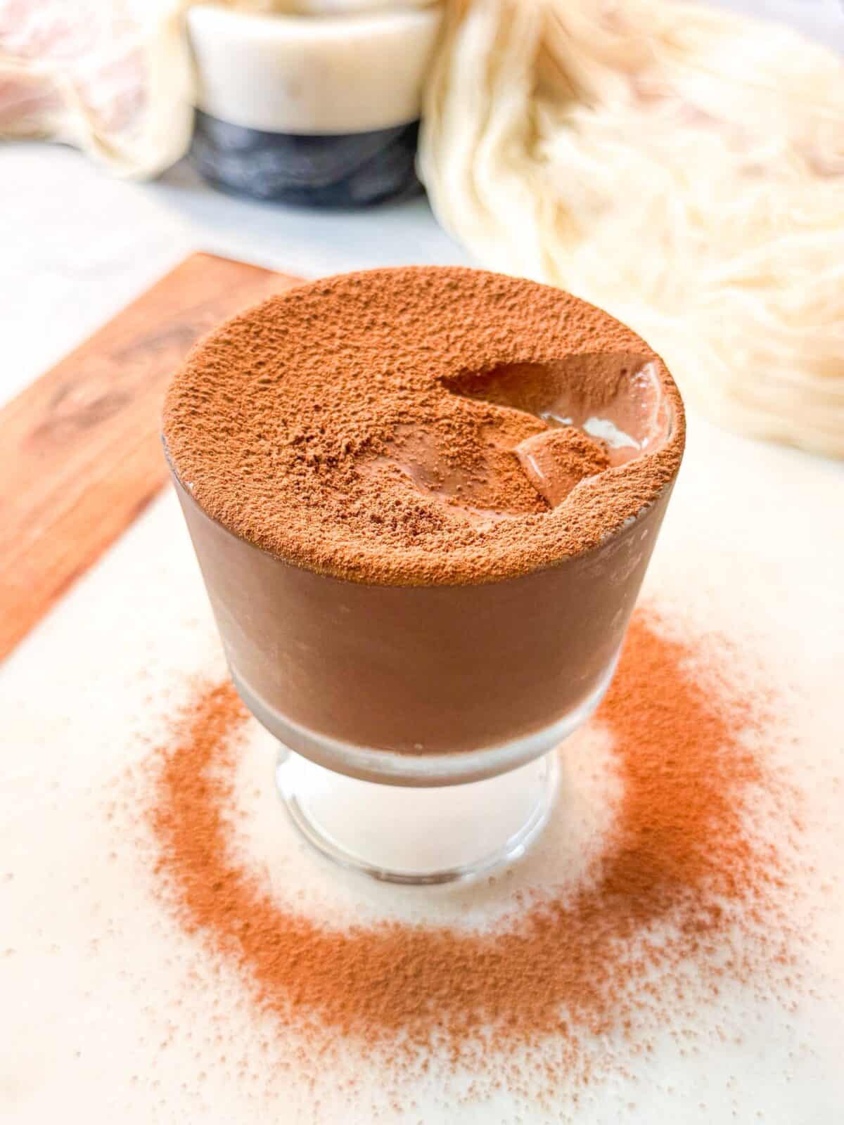 A perfectly smooth chocolate pudding served in a glass, topped evenly with a layer of fine cacao powder.