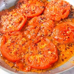 close-up shot of juicy garlic-lemon roasted tomato slices in a skillet with flavorful pan juices, and sprinkled with crushed red pepper flakes