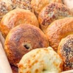 close-up of freshly baked homemade bagels with a variety of toppings such as melted cheese and sesame seeds, placed on parchment paper in a metal tray.