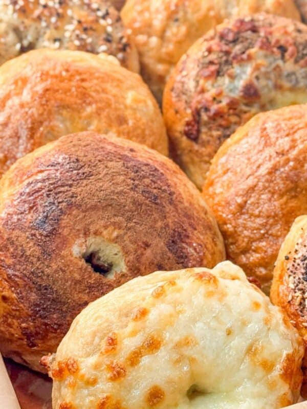 close-up of freshly baked homemade bagels with a variety of toppings such as melted cheese and sesame seeds, placed on parchment paper in a metal tray.