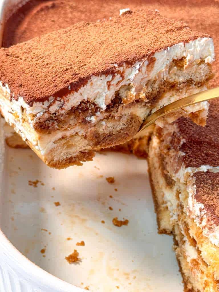 Best Eggless Tiramisu Recipe a piece of eggless tiramisu is being lifted out of the baking dish with a gold spoon, highlighting the creamy layers and cocoa-dusted top