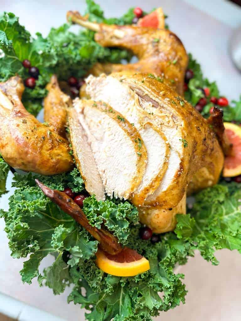Best Homemade Thanksgiving Turkey close-up shot of a juicy roasted turkey with several breast slices carved, served on a bed of fresh kale, cranberries, and orange slices