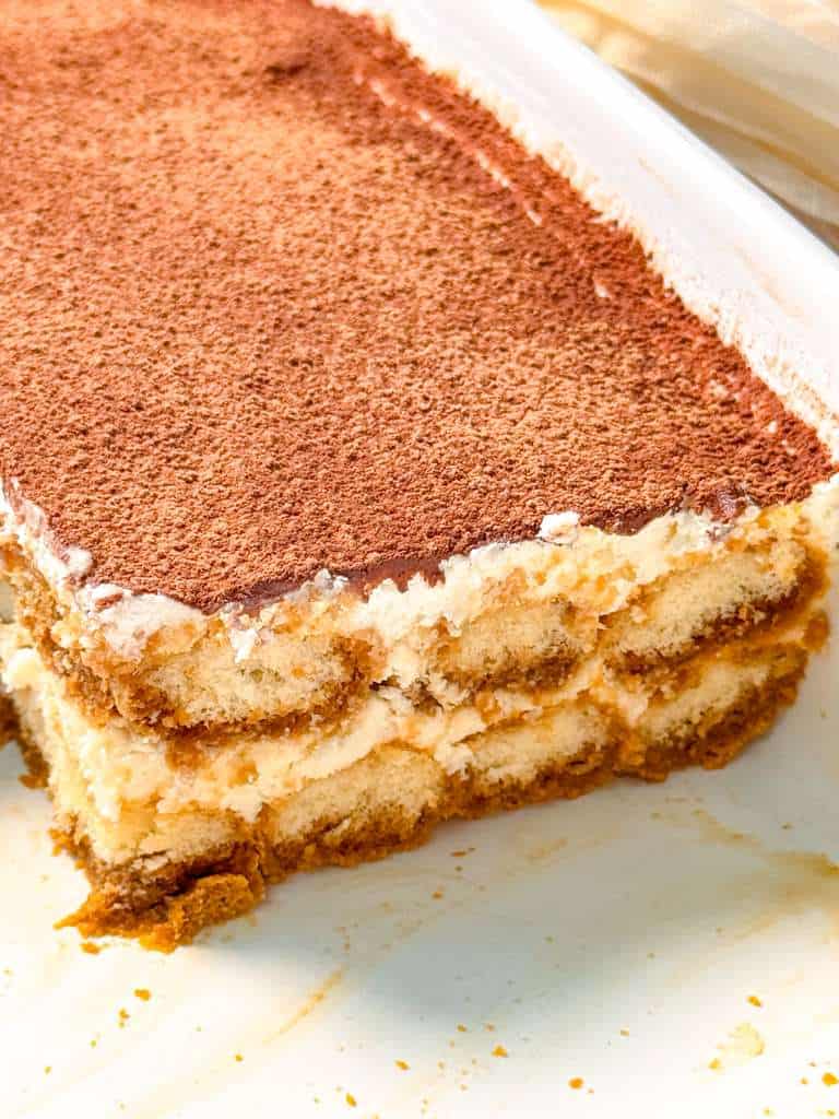 Easy Eggless Tiramisu Recipe a close up shot of a slice of eggless tiramisu on a white plate, displaying its distinct layers of cream, soaked ladyfingers, and a thick dusting of cocoa powder on top