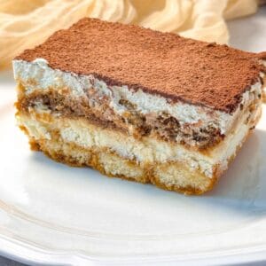 a slice of eggless tiramisu on a white plate, displaying its distinct layers of cream, soaked ladyfingers, and a thick dusting of cocoa powder on top