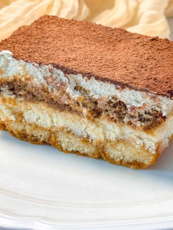 a slice of eggless tiramisu on a white plate, displaying its distinct layers of cream, soaked ladyfingers, and a thick dusting of cocoa powder on top