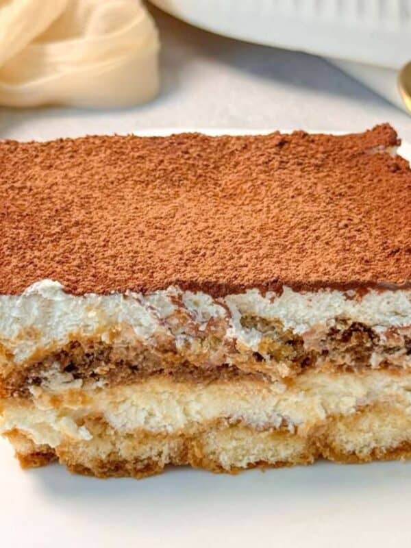 a slice of eggless tiramisu on a white plate, displaying its distinct layers of cream, soaked ladyfingers, and a thick dusting of cocoa powder on top