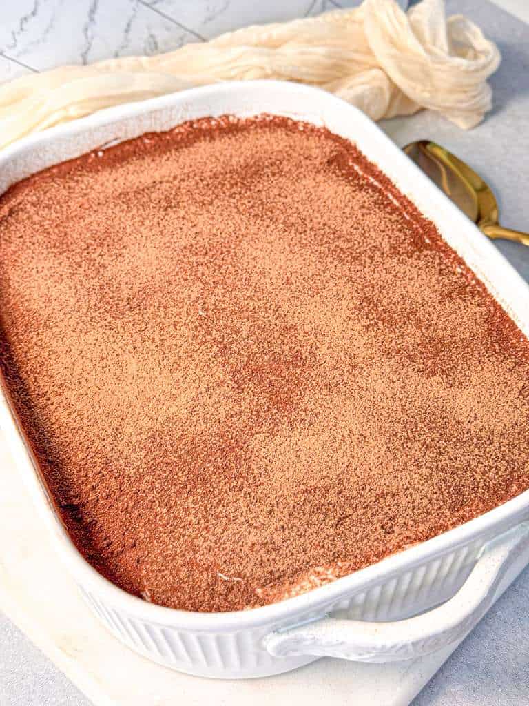 Tiramisu Egg Free eggless tiramisu served in a white baking dish, showing layered espresso-soaked ladyfingers, creamy mascarpone filling, and a smooth cocoa powder topping