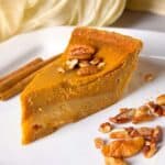 a close shot of a slice of pumpkin pie topped with pecans on a white plate, served beside cinnamon sticks and chopped nuts