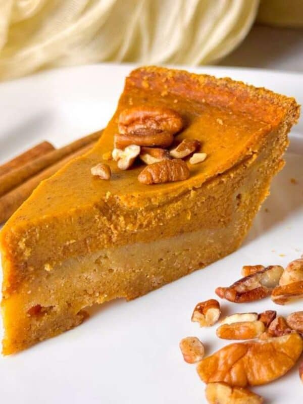 a close shot of a slice of pumpkin pie topped with pecans on a white plate, served beside cinnamon sticks and chopped nuts