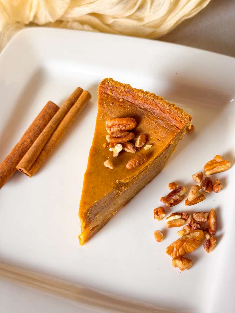 Homemade Pumpkin Pie Recipe a top shot of a slice of classic homemade pumpkin pie topped with pecans on a white plate, served beside cinnamon sticks and chopped nuts