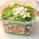 A close-up shot of a meal prep layered salad in a square glass meal prep container, made with arugula, sprouts, and crumbled feta cheese, displayed on a kitchen counter.