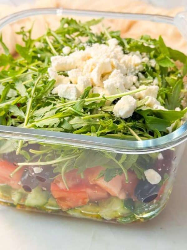 A close-up shot of a meal prep layered salad in a square glass meal prep container, made with arugula, sprouts, and crumbled feta cheese, displayed on a kitchen counter.