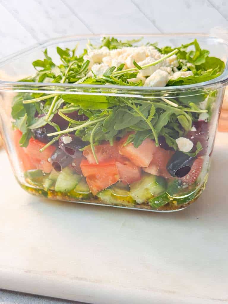 Meal-Prep Layered Salad Recipe A close side shot of a layered meal prep salad in a glass container showing cucumbers, tomatoes, olives, dressing at the bottom, and arugula with feta on top.