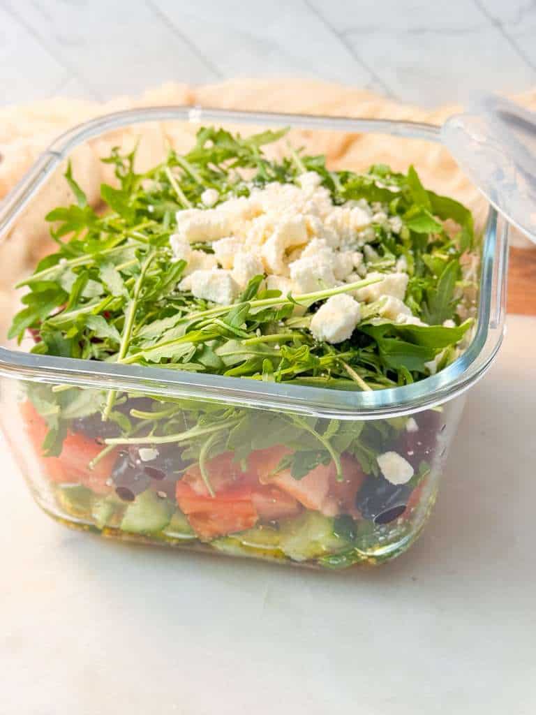 No Soggy Meal-Prep Salad A shot of meal prep layered salad in a square glass meal prep container made with arugula, sprouts, and crumbled feta cheese, displayed on a kitchen counter.