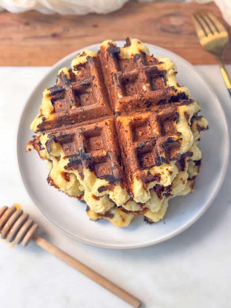 High Protein Cottage Cheese Waffles Top shot of fluffy cottage cheese waffles stacked on a small plate, served on a marble board with a honey dipper and fork beside them.