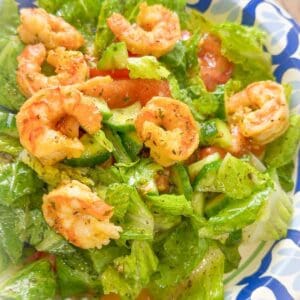Close-up of cooked shrimp served on top of salad with romaine lettuce, cucumbers, and tomatoes, tossed in dressing and served in a patterned bowl.