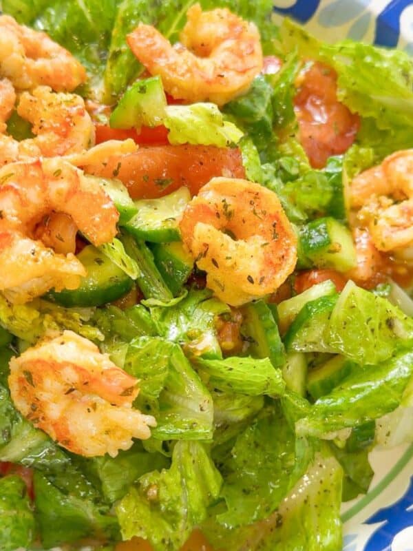 Close-up of cooked shrimp served on top of salad with romaine lettuce, cucumbers, and tomatoes, tossed in dressing and served in a patterned bowl.