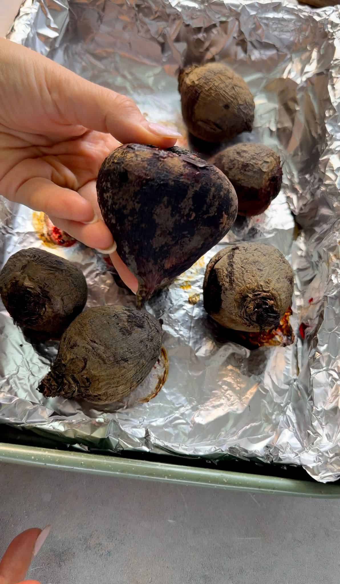 Steps to Roast Beets Hand holding a roasted beet over a foil-lined baking dish filled with whole roasted beets.