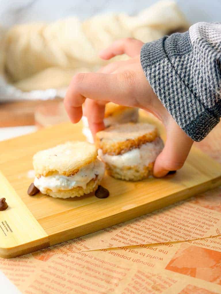 Air Fryer Crispy Calrose Rice Ice Cream Sandwiches Hand placing crispy rice ice cream sandwiches with vanilla filling on a wooden board, with chocolate chips scattered around.