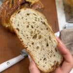 close shot of a woman holding a slice of easy overnight artisan sourdough bread with an airy open crumb and crisp golden crust, with the whole loaf in the background