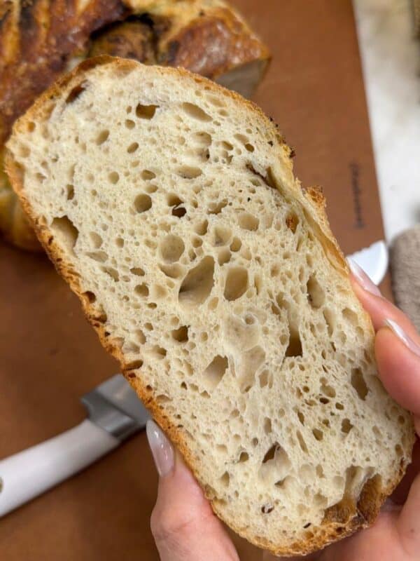 close shot of a woman holding a slice of easy overnight artisan sourdough bread with an airy open crumb and crisp golden crust, with the whole loaf in the background